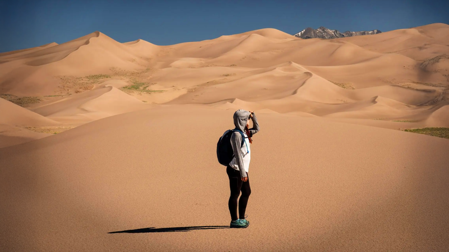 Person with a backpack stands on desert sand dunes under clear blue sky.