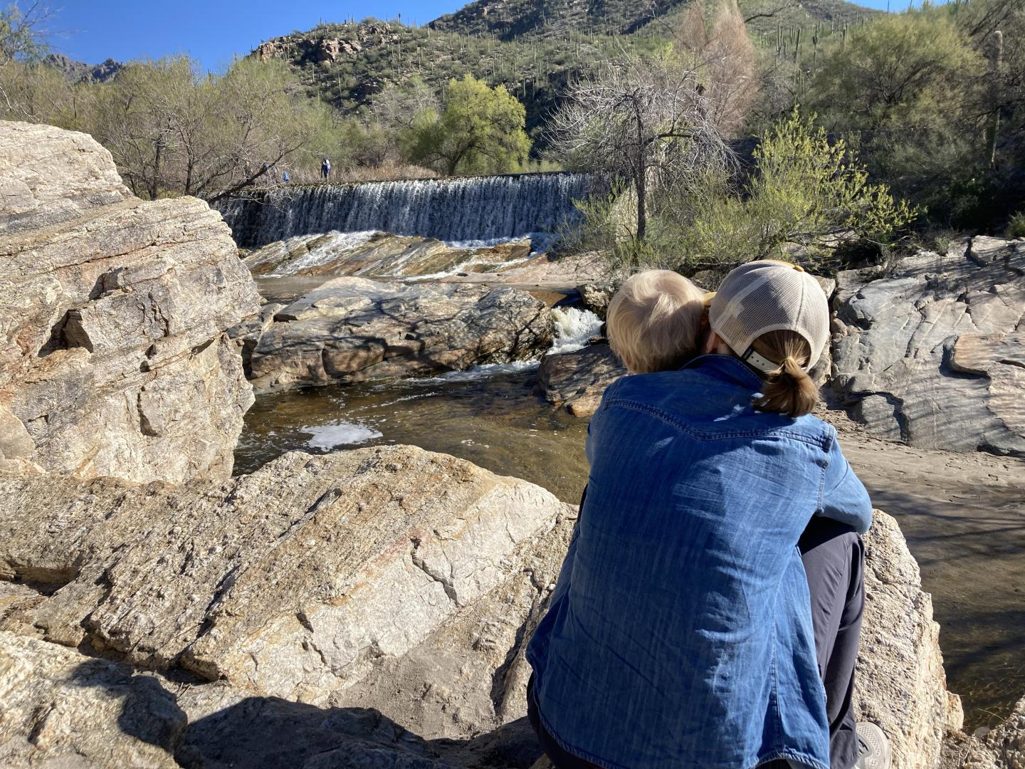 Person sitting on rocks by a stream, embracing a child.