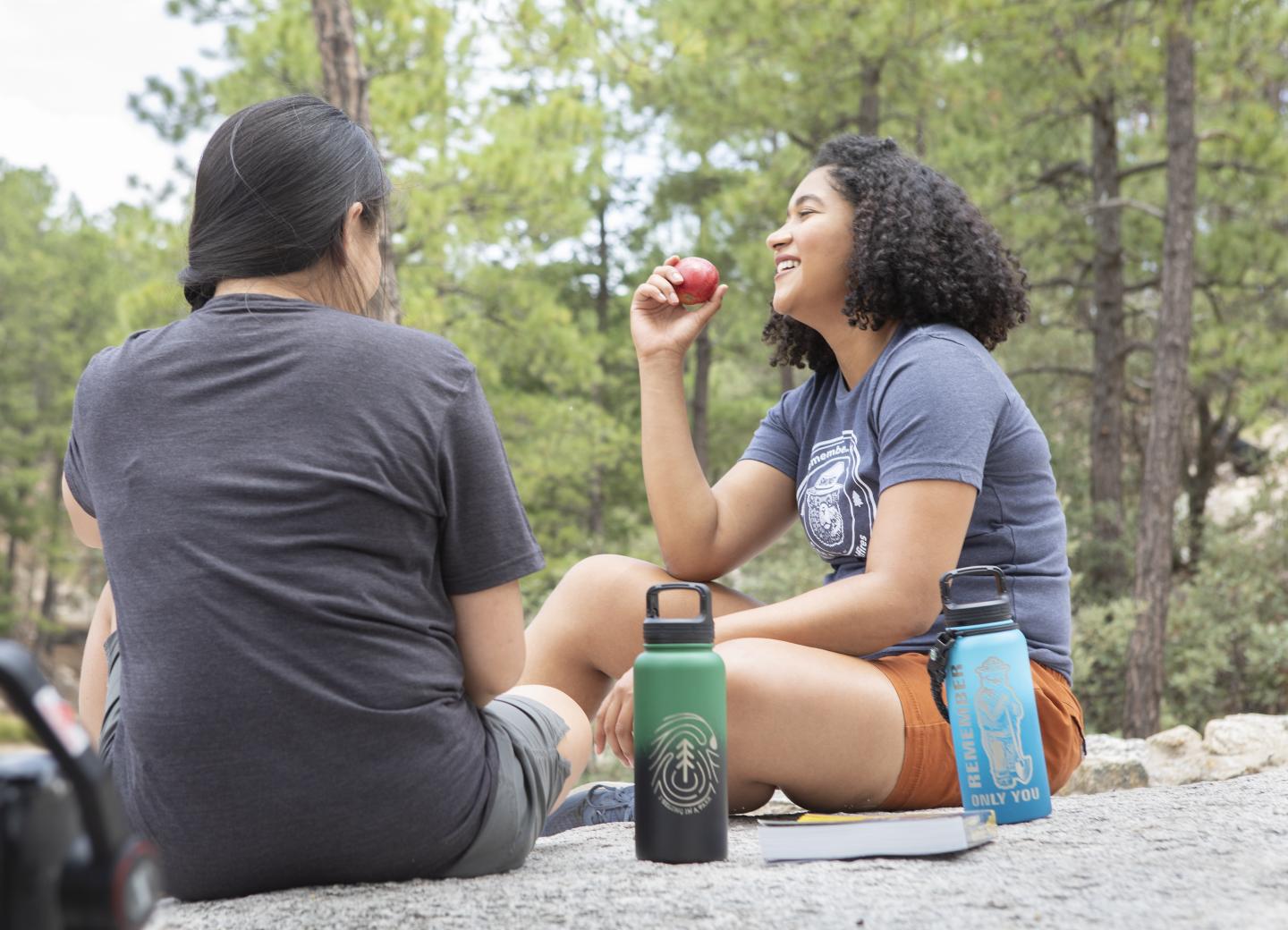 Two people picnicking on a rock in Mount Lemmon, chatting and eating, with trees in the background.