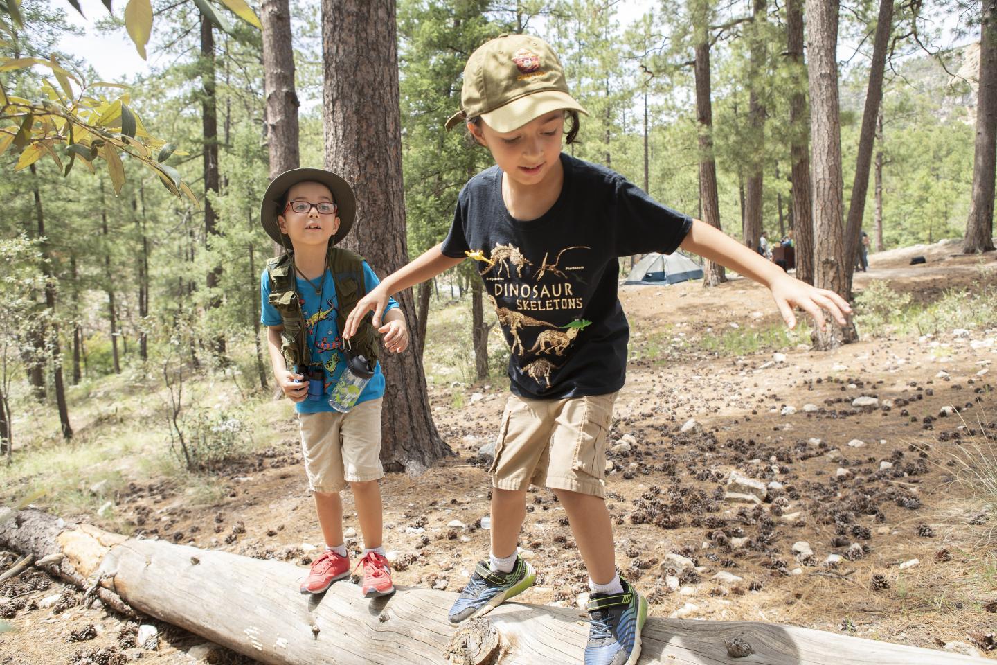 Two kids balancing on a log in a forest, wearing hats and casual clothes.