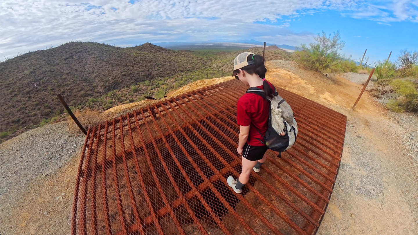 Hiker in red shirt and cap stands on rusted grate, desert landscape in background.