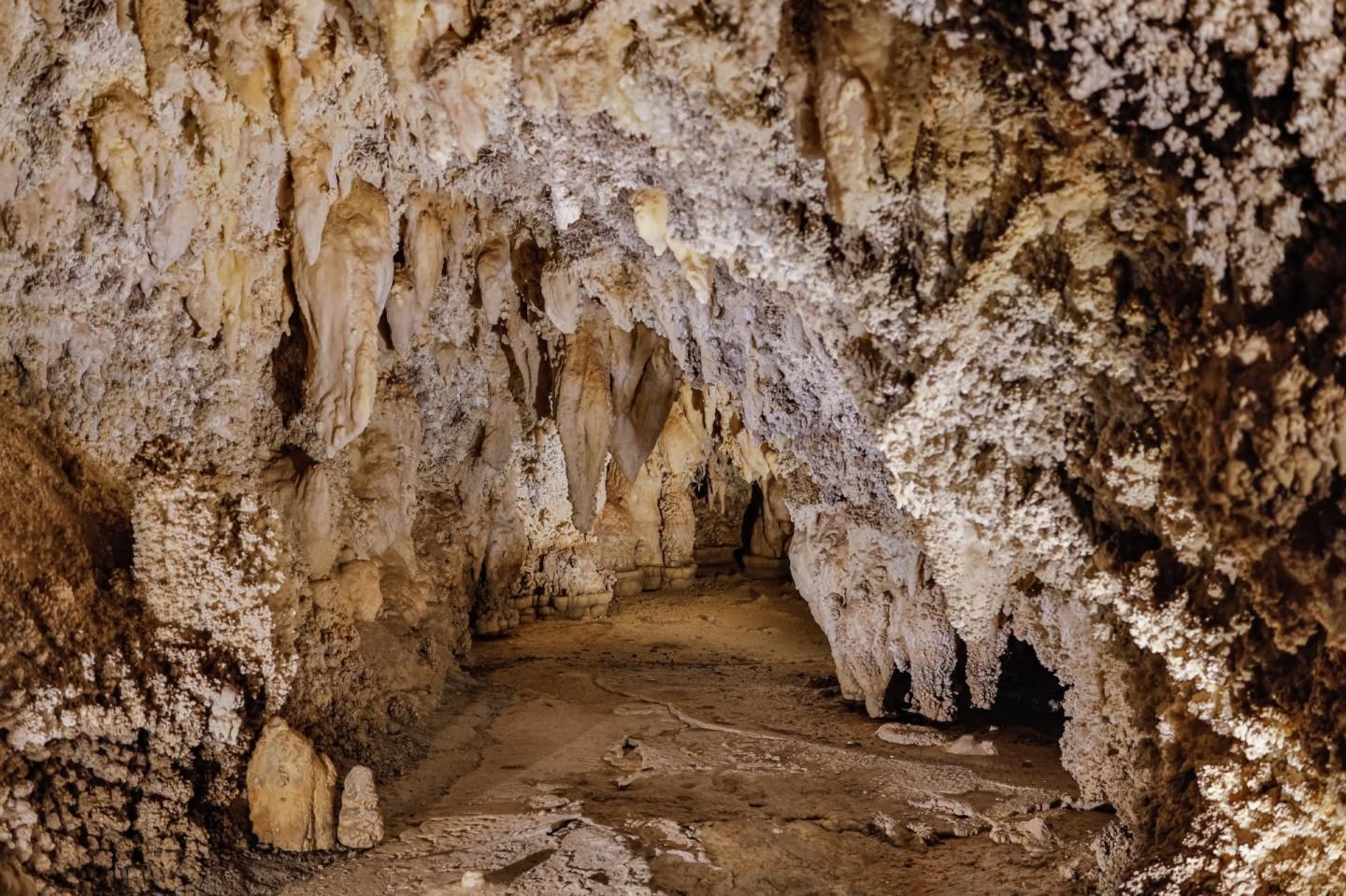 Cave interior with stalactites and stalagmites, dimly lit with a sandy floor.