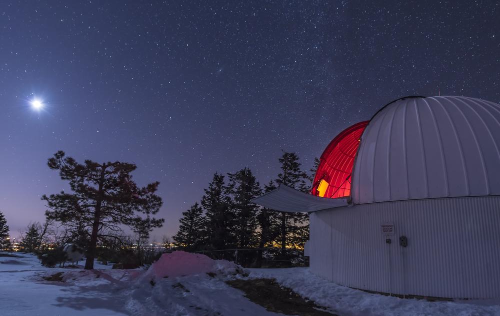 Observatory under starry sky with a bright moon and surrounding trees.