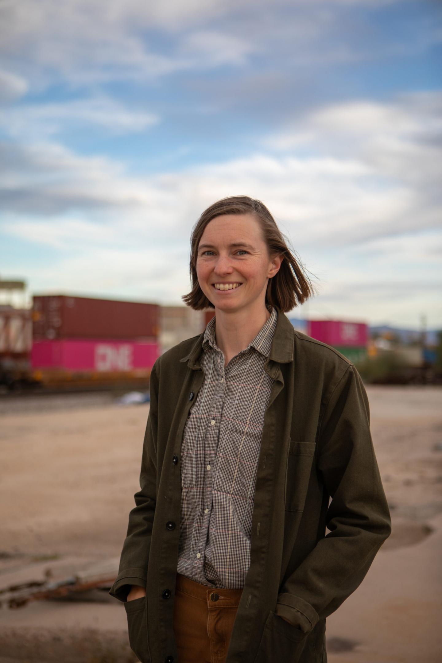 Author Caroline Tracey smiling, wearing a jacket standing infront of colorful shipping containers under a blue sky.