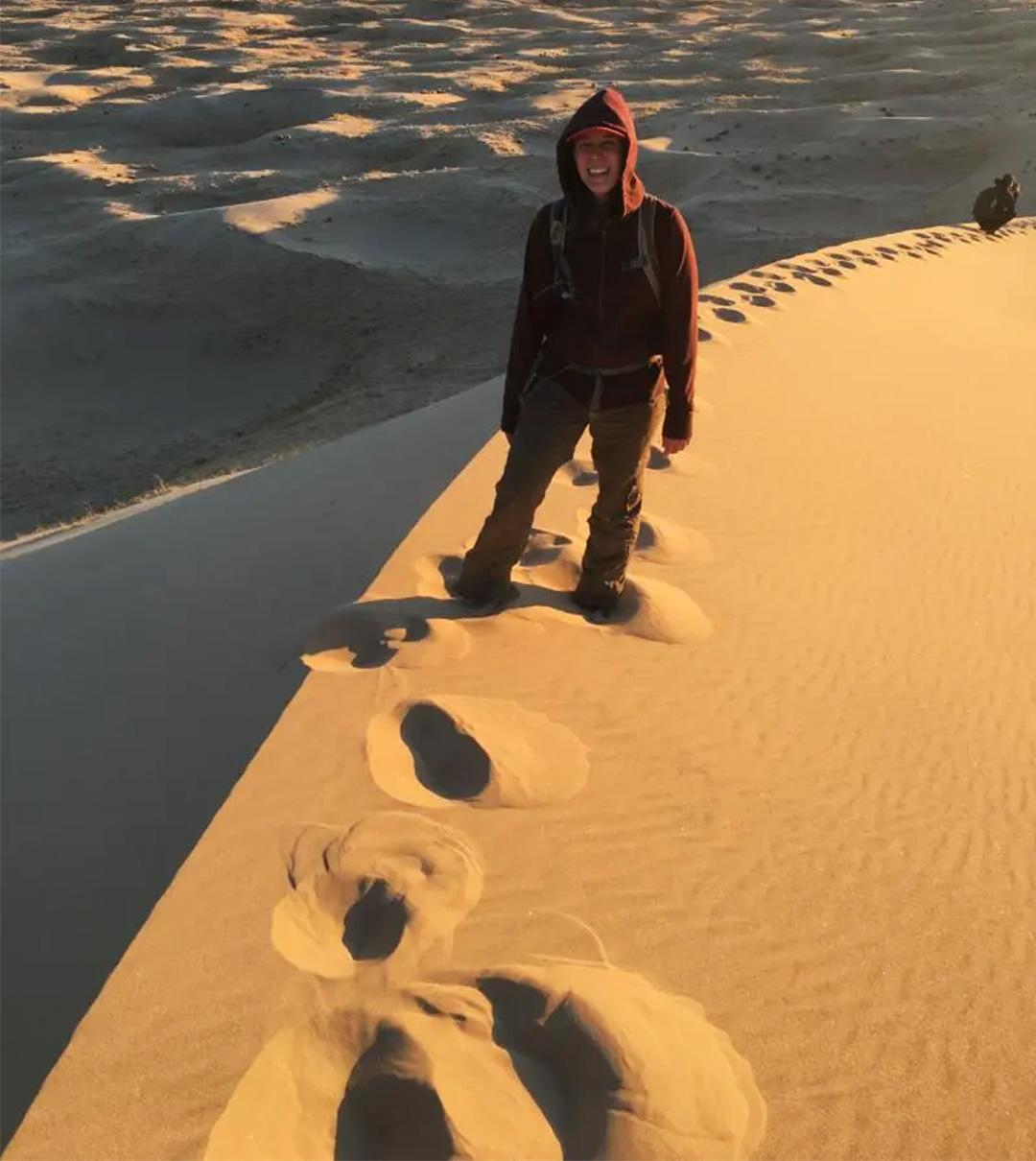 Person in a hoodie standing on sunlit sand dune, with footprints behind them.