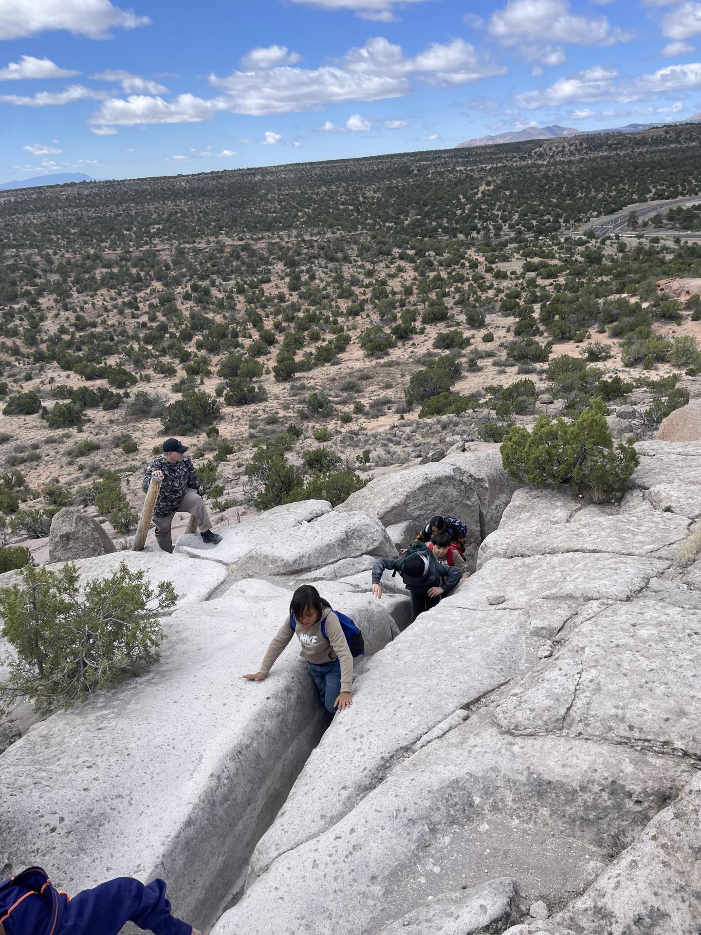 Young hikers ascending a crevice on a rocky hill, with vast green landscape and cloudy sky.