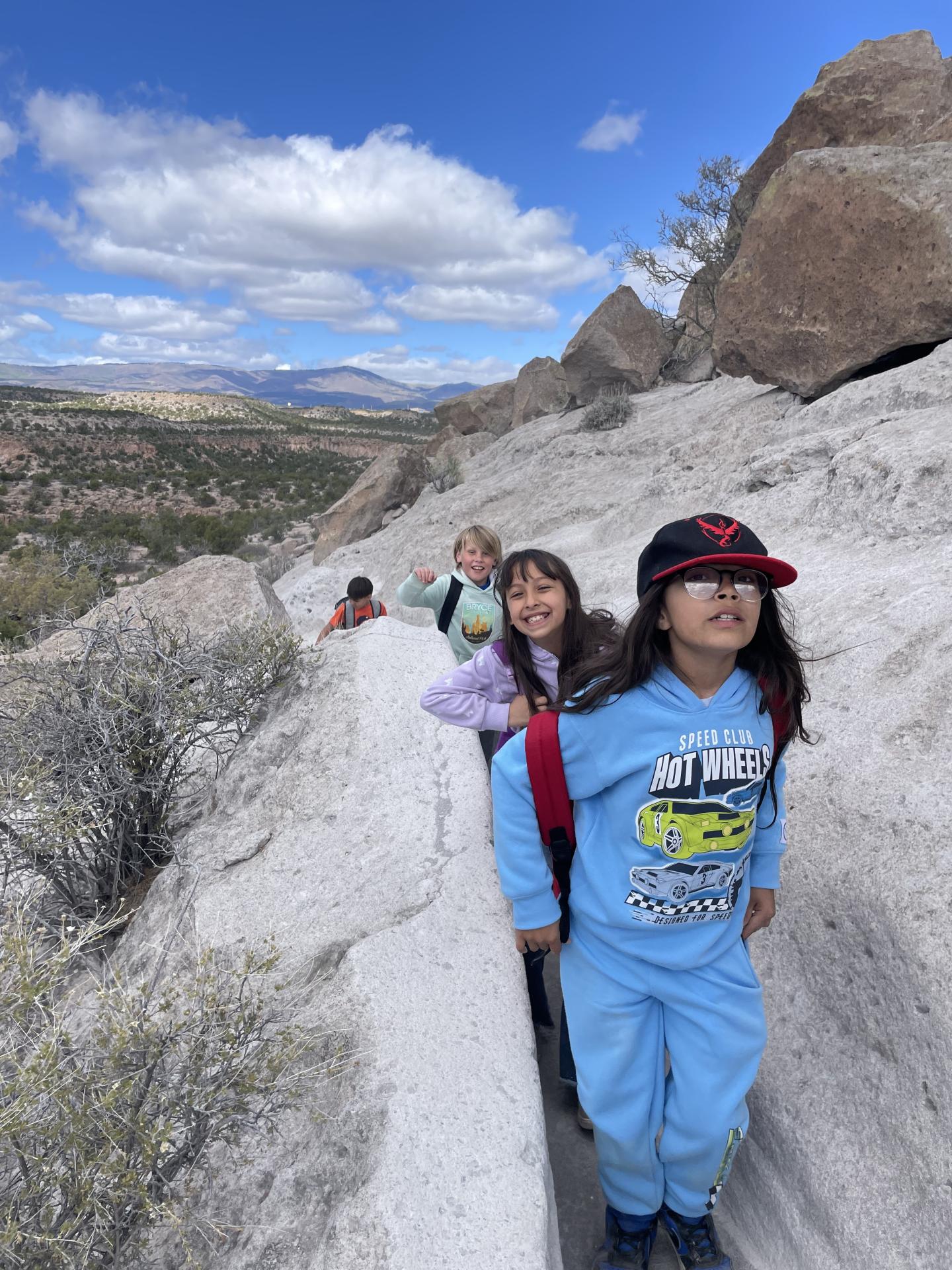 Children hiking on a rocky trail under a blue sky with clouds.