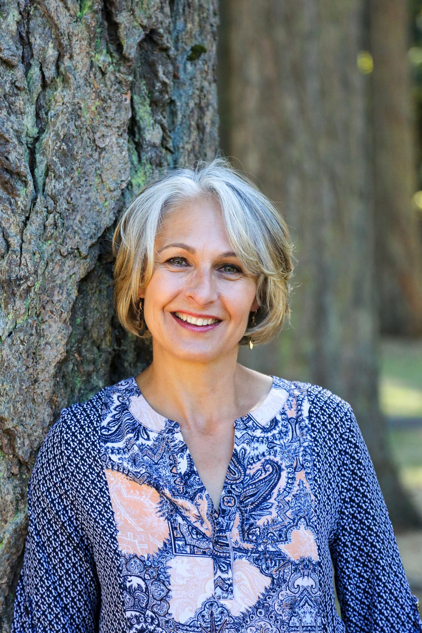 Woman with short gray hair smiling by a tree in a patterned blue blouse.