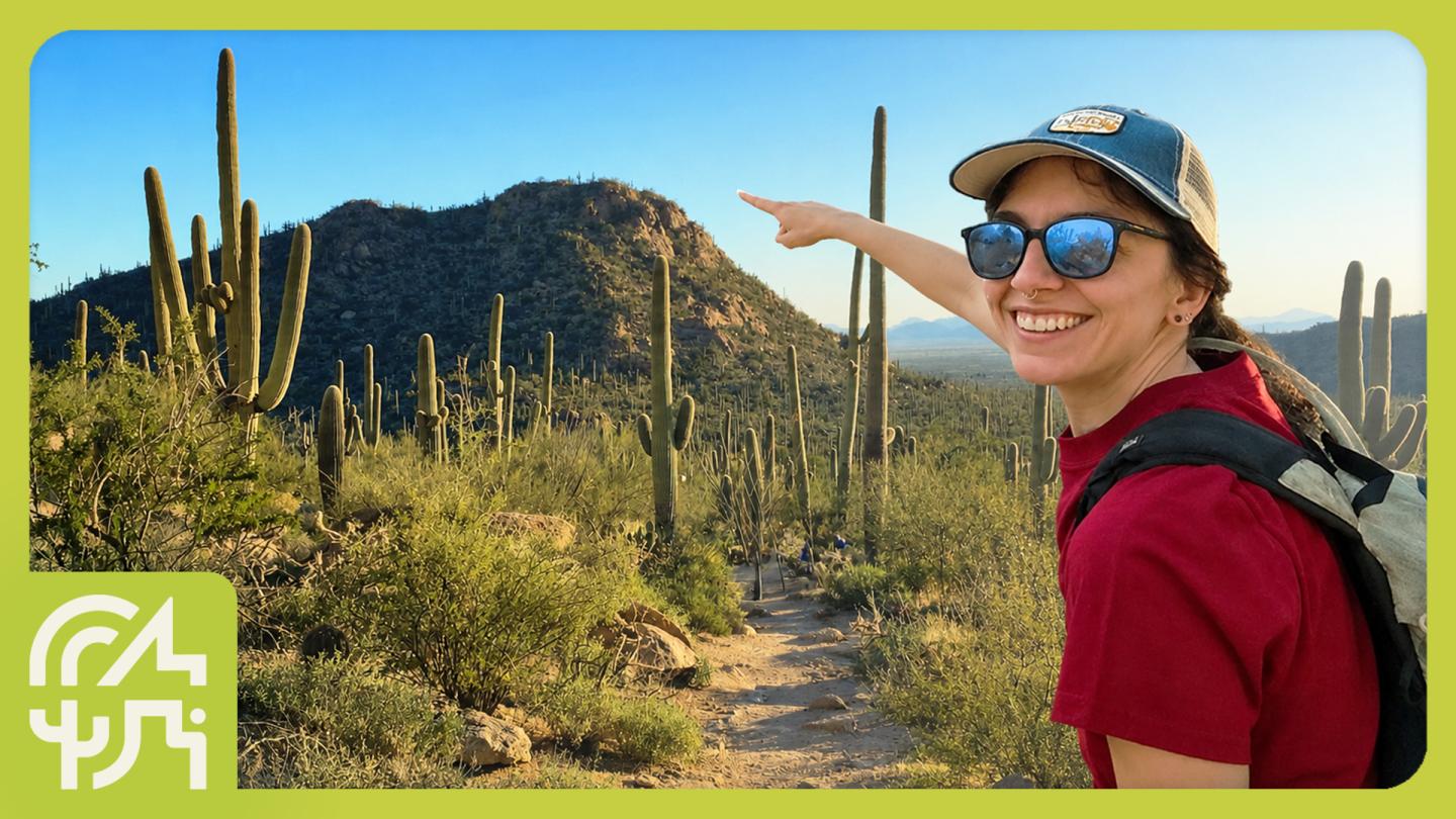 Woman in desert pointing at mountain, wearing a hat and sunglasses, smiling.