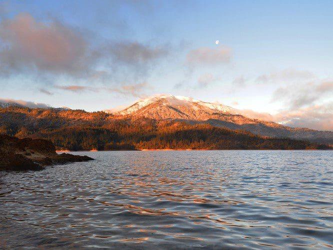 A snowy mountain at sunset at Whiskeytown National Recreation Area. The moon is visible in the sky. The foreground is the calm waters of Whiskeytown Lake.