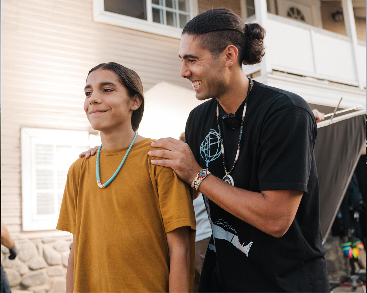 Two people smiling outside a house, one with hands on the other's shoulders.