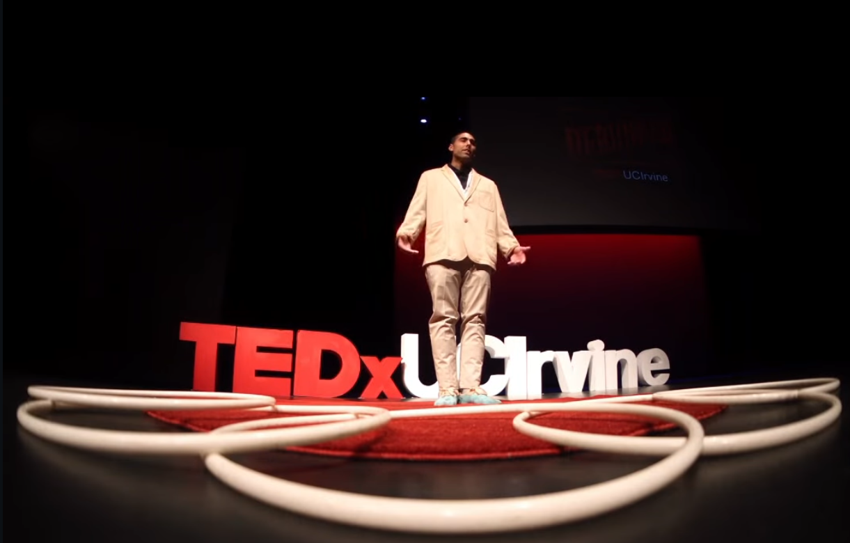 Man speaking at a TEDx event, wearing a light suit, red stage lighting.