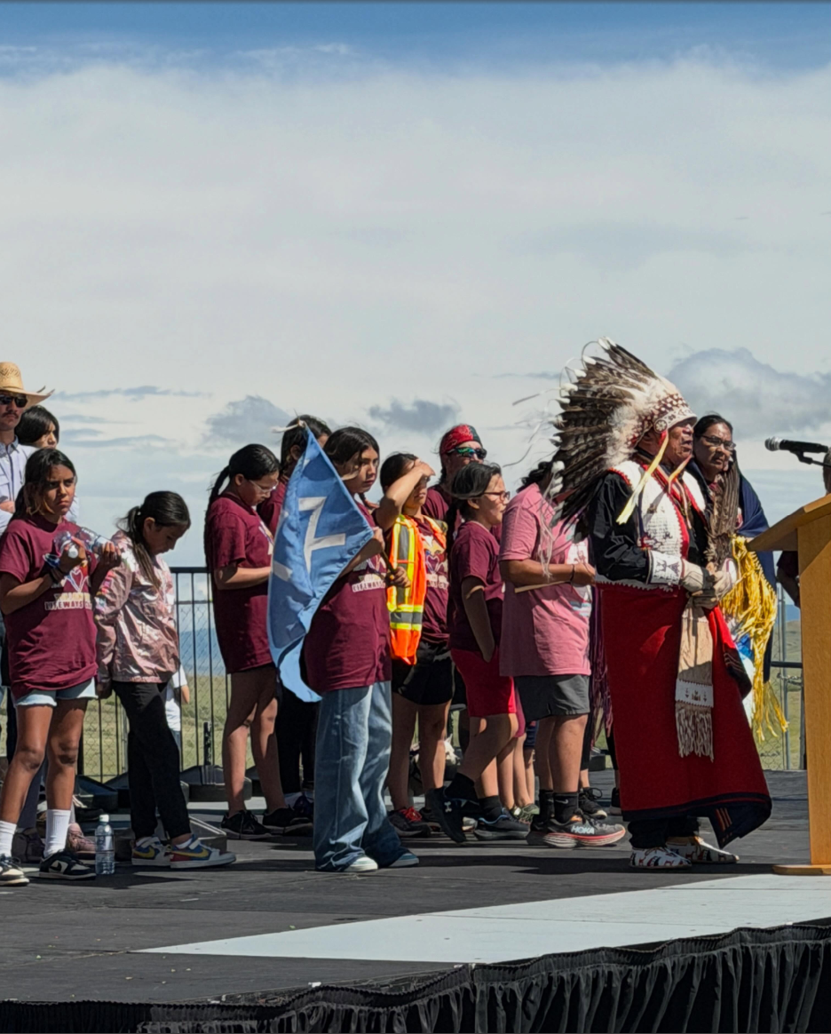 People on a stage, one wearing a headdress, holding a blue flag, under a cloudy sky.