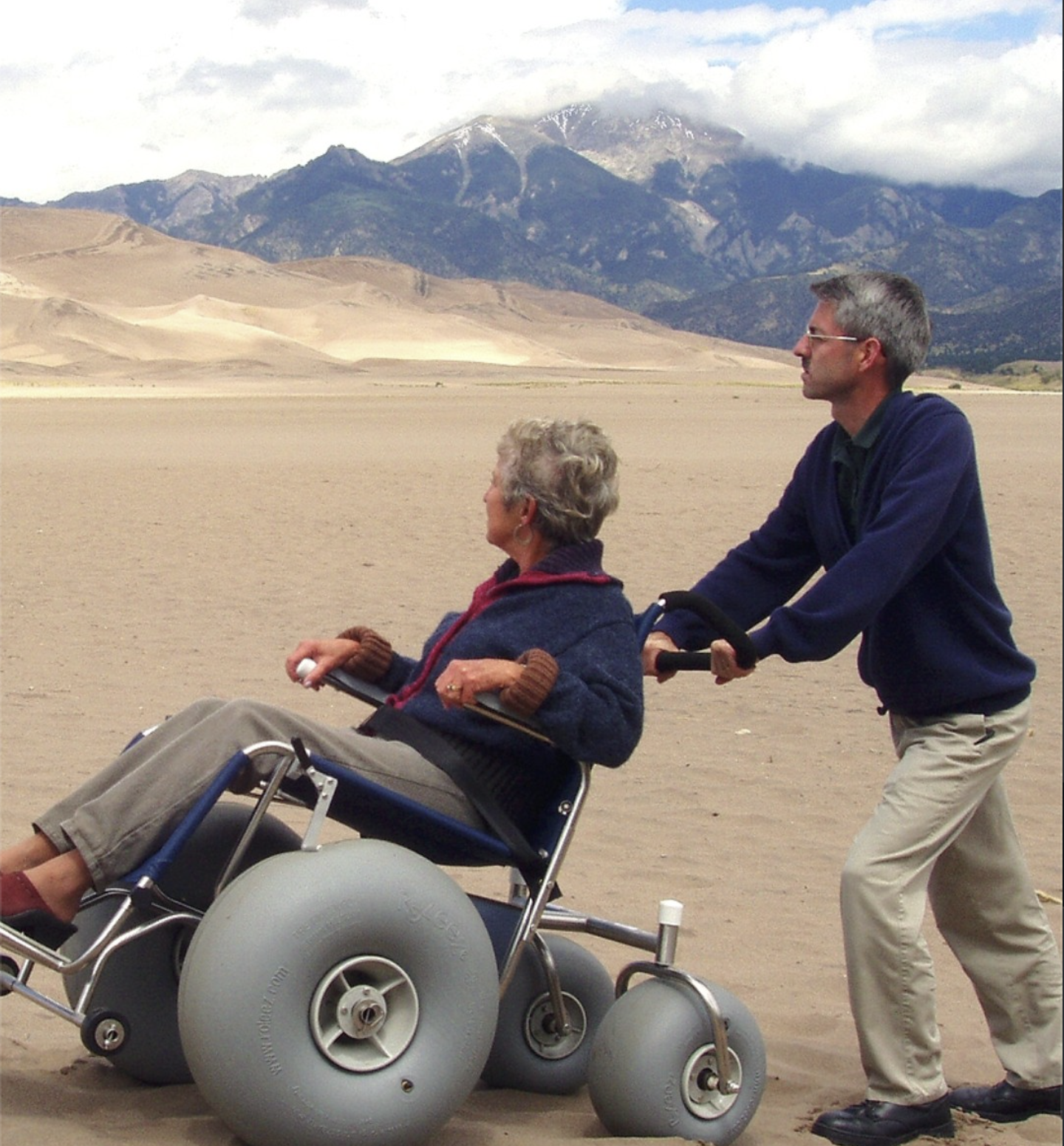 Man pushing woman in wheelchair on sandy terrain with mountains in background.