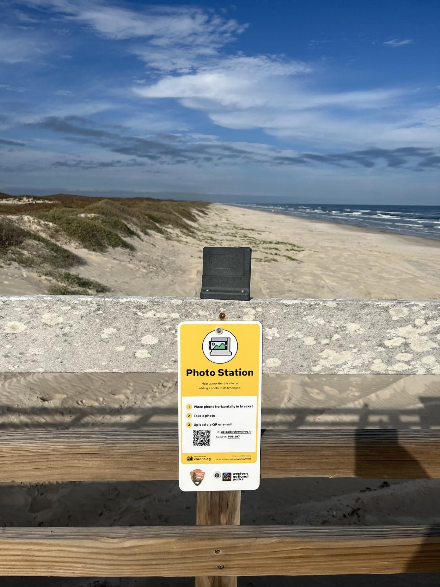 Beach scene with a sand path, ocean waves, and a phone holder on a wooden railing.
