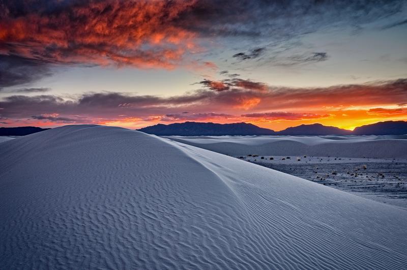 Desert landscape at sunset with orange and purple sky over sand dunes.