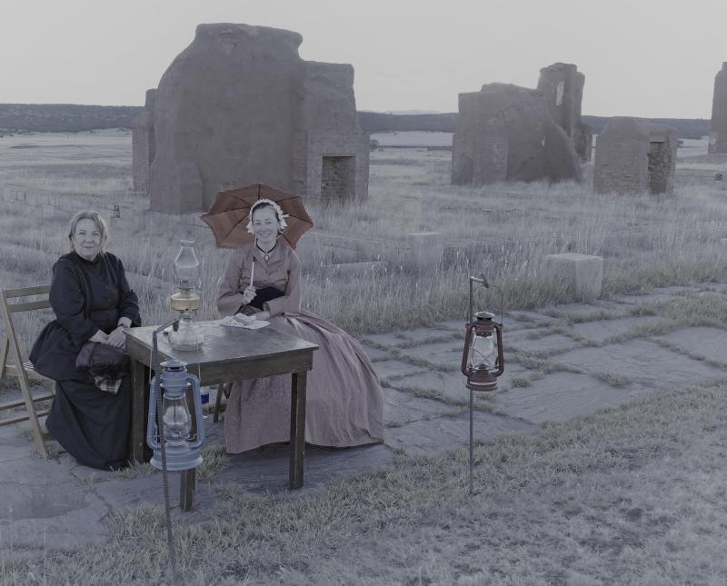Two seated costumed living history interpreters near adobe ruins.