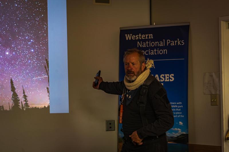 A man stands indoors pointing at an obscured projected image of a computer screen behind him