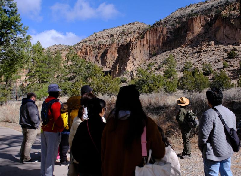 a group of people stand facing a ranger in a green uniform looking up towards a cliff face.
