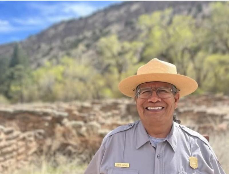a smiling ranger in a wide brimmed hat stands in front of a historic small rock wall.