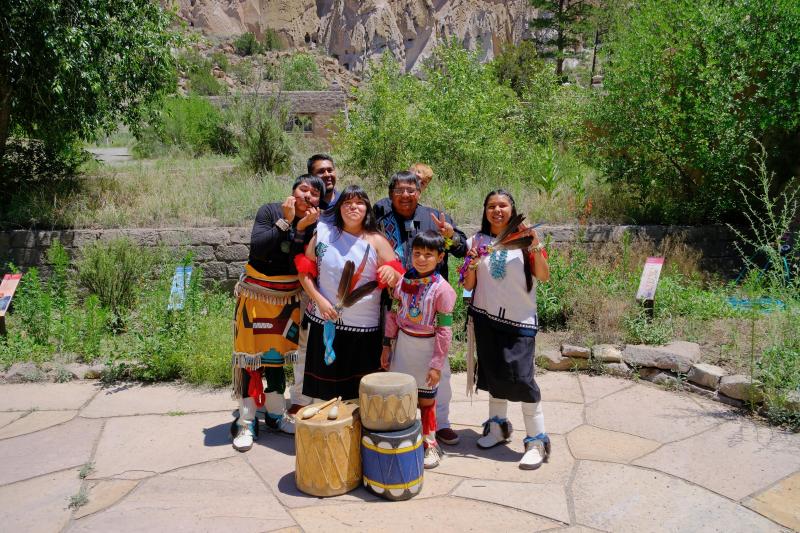 a pueblo group stands smiling in colorfol regalia and drums at their feet in front of the canyon wal