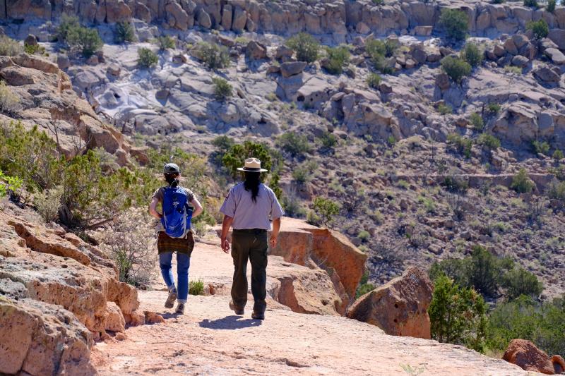 Ranger and park visitor walk along the rim of a mesa made of orange volcanic tuff.