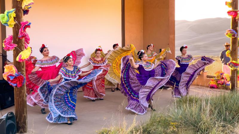 A group of 7 women dancers in colorful dresses dancing on stage with sand dunes in the background
