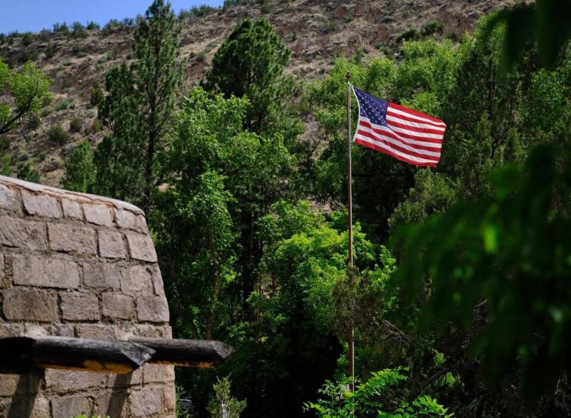 An American flag on a flagpole waves in the wind in front of the corner of a stone masonry building.
