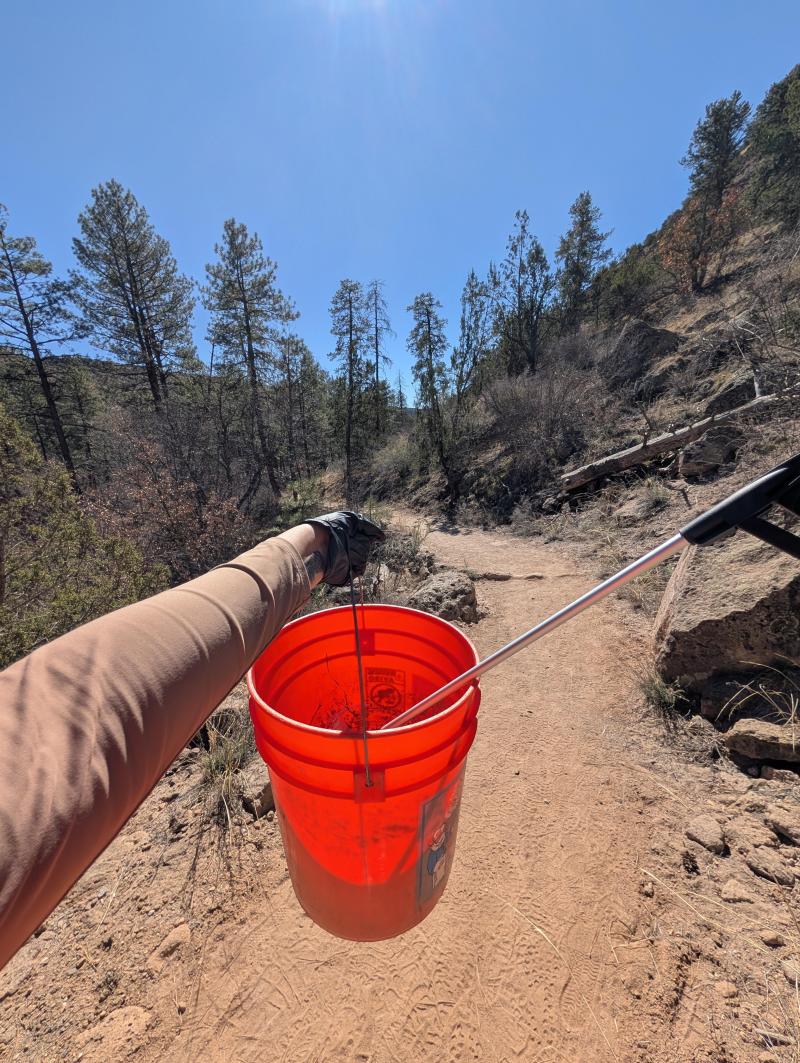 A point of view of an arm outstretched, holding an orange bucket with a litter grabber on a trail.