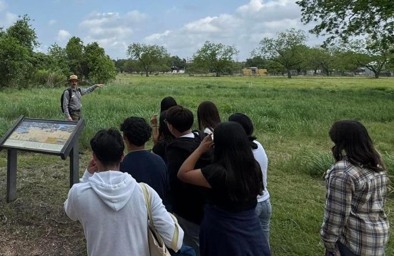 A park ranger gestures to a large grassy field while a group of people give their attention to him.