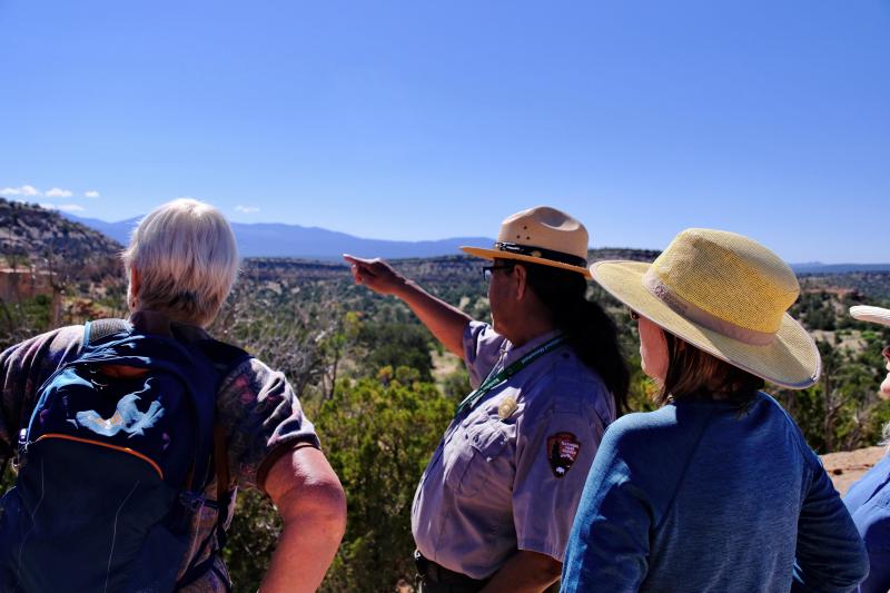 A ranger standing with two park visitors on a desert cliffside points to mountains in the distance.