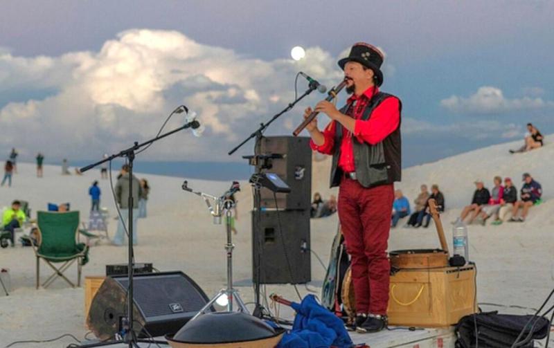 A man in a red shirt plays a flute in front of an audience sitting in white sand dunes