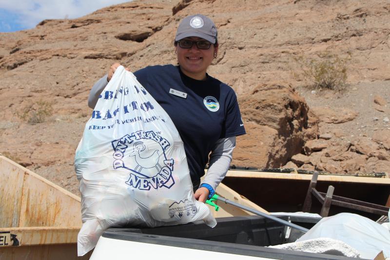 Individual holding a full trash bag in front of a dumpster