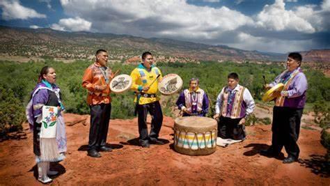 Black Eagle Singers pictured on a red sandstone ledge with the Jemez mountains in the background.