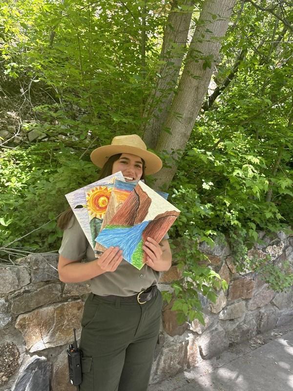 A woman park ranger in a forest holds up pieces of hand-drawn art in front of her.