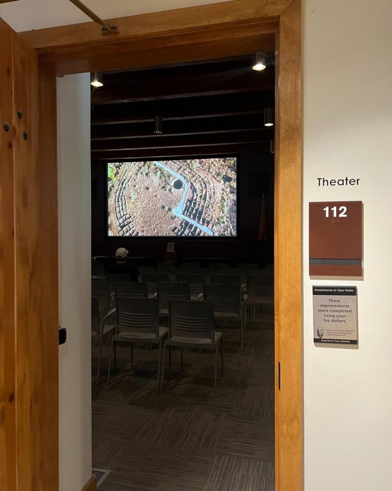 A visitor sits in a dark room with a large projector screen showing an image of a Pueblo site.