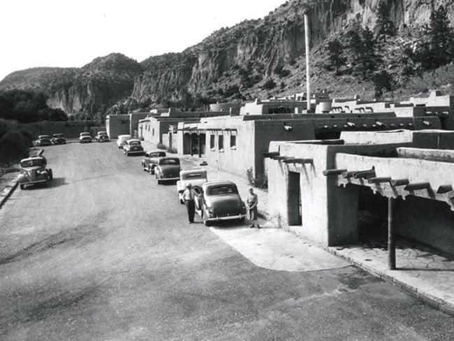 Black and white photo of visitors exiting car parked alongside row of masonry buildings at the base