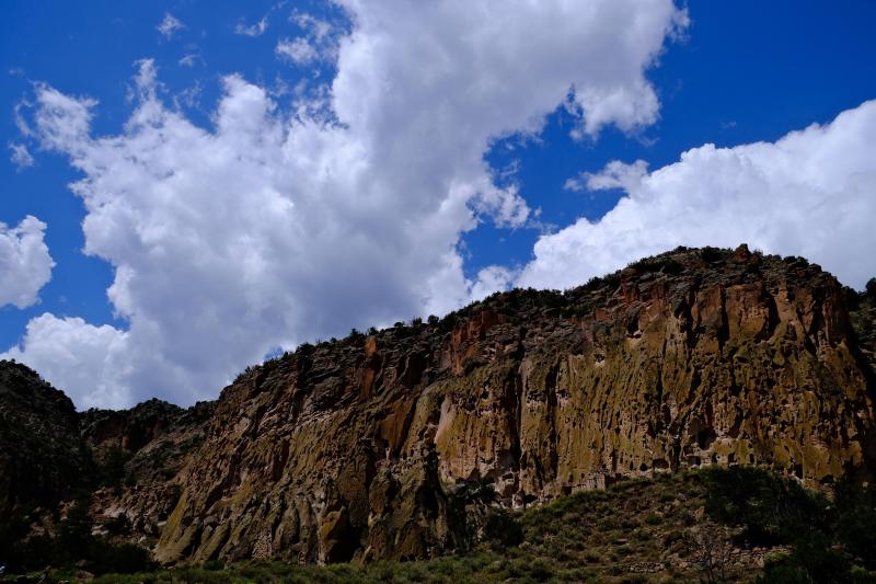 Large, white clouds fly across bright, blue skies over a light orange cliffside.