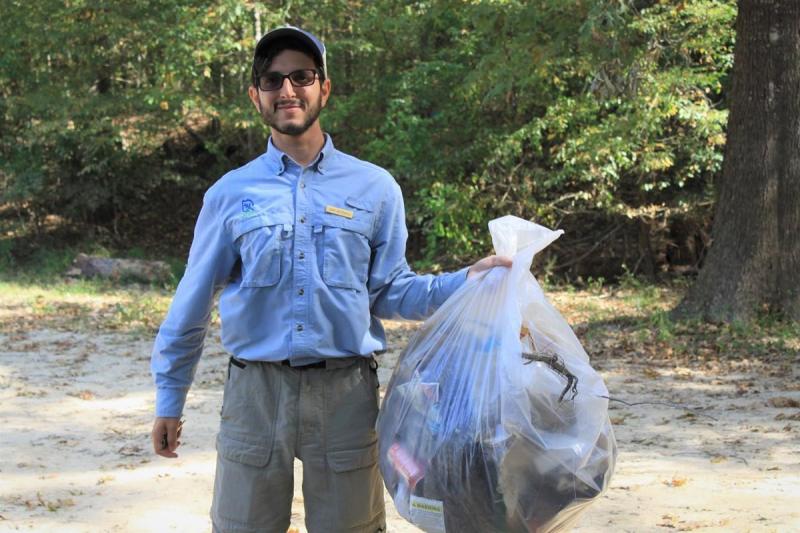 A man wearing blue shirt and ball cap holding a full bag of trash outside a forest.