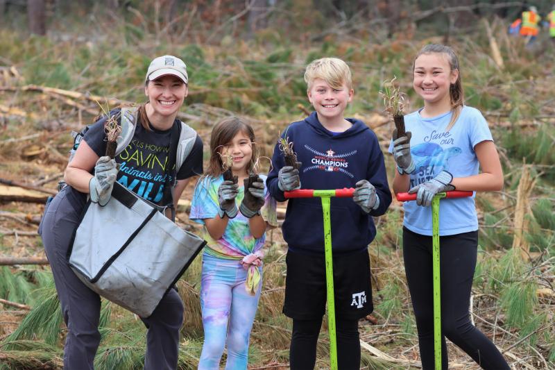 A smiling family, with a mother and 3 kids, holding green dibble bars and a bag of plant seedlings.