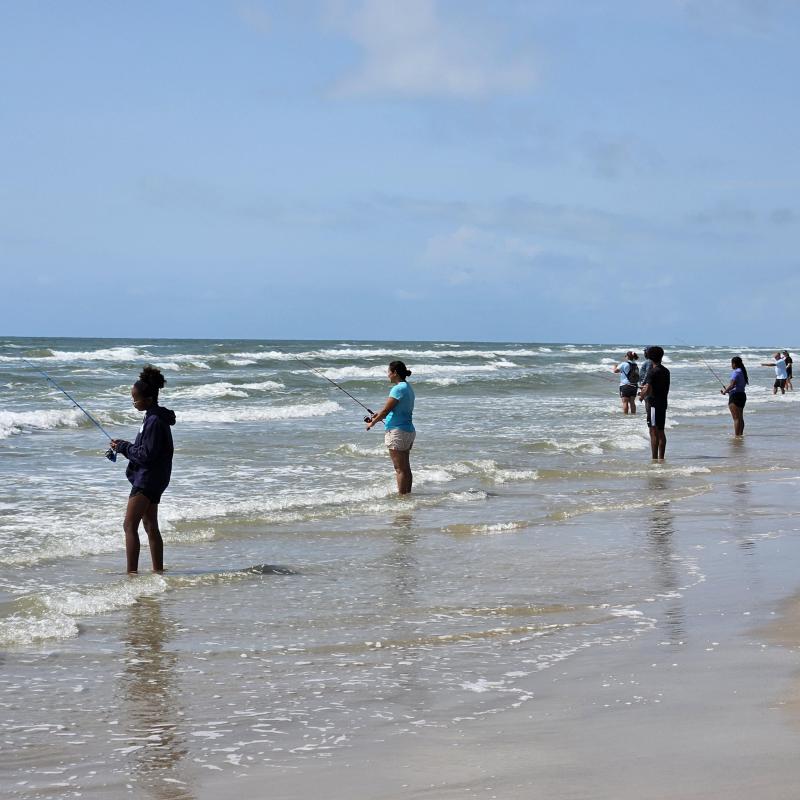 A group of people standing on the edge of the beach while fishing.
