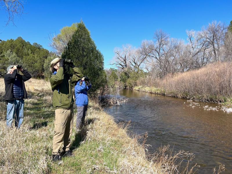 Three people stand next to a river, each looking through binoculars.