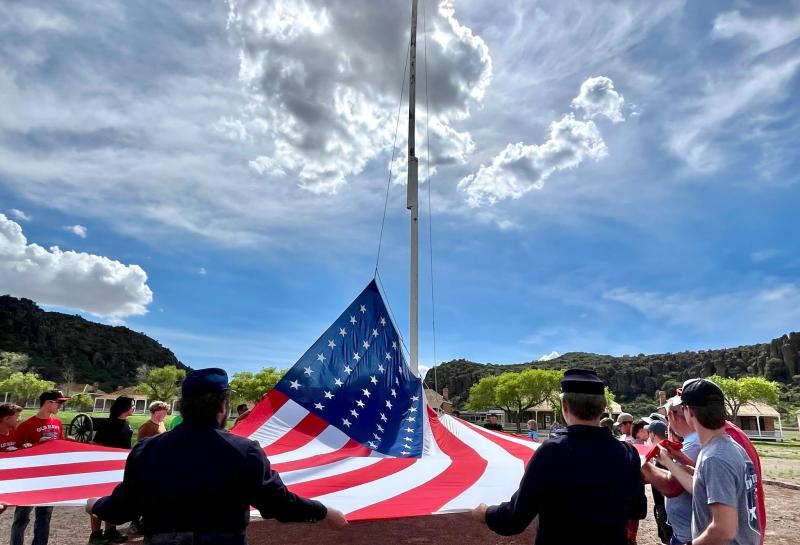 People hold a large red, white, and blue flag around the edges at the base of a flag pole