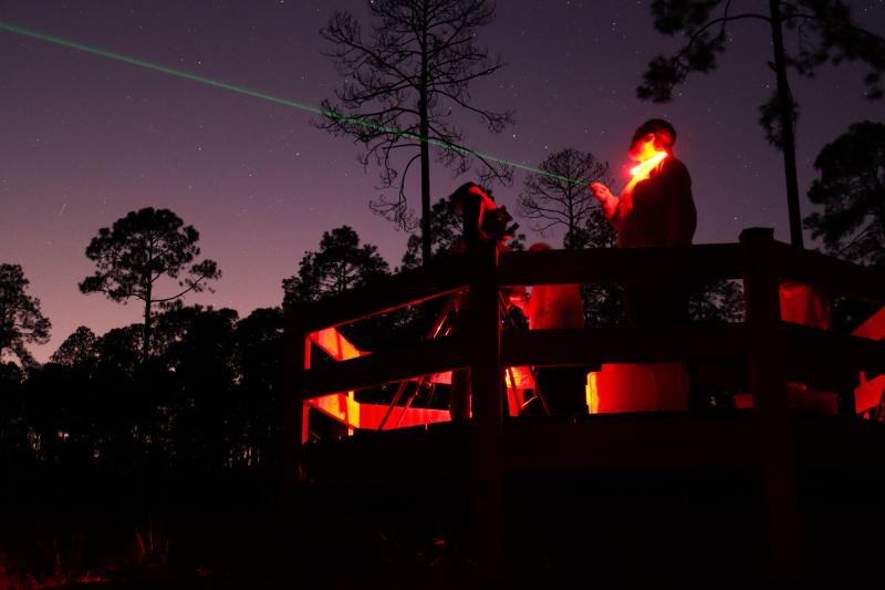 Nighttime photo of a man pointing a green laser to the night sky with red lights illuminating him.