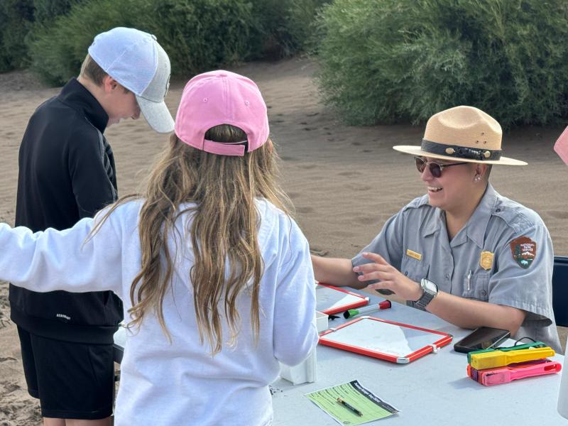 A Park ranger with flat hat sits at a table with two children observing black sand and magnets