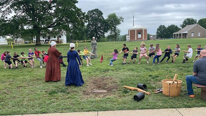 Children in a tug-of-war game on a grassy field with 3 women in historical dresses observing nearby.