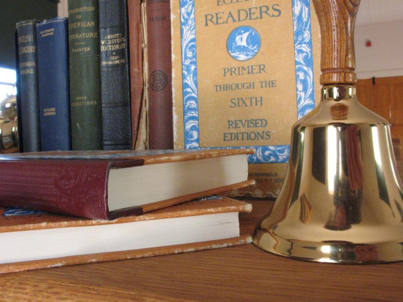 A brass handbell stands next to a stack of early 20th century schoolbooks.