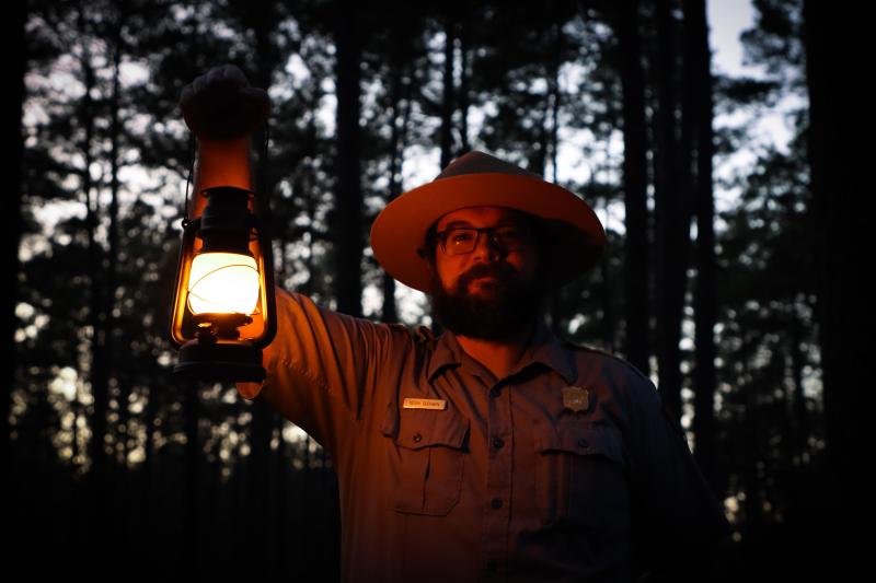 A park ranger illuminated by the vintage lantern he is holding up in his right hand.