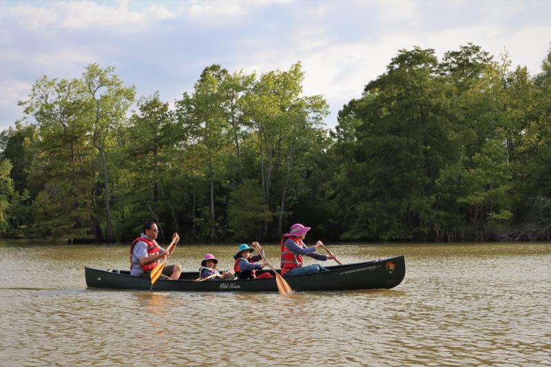 family of 4, including a father, 2 daughters, and a mother, paddling a green canoe on a river.