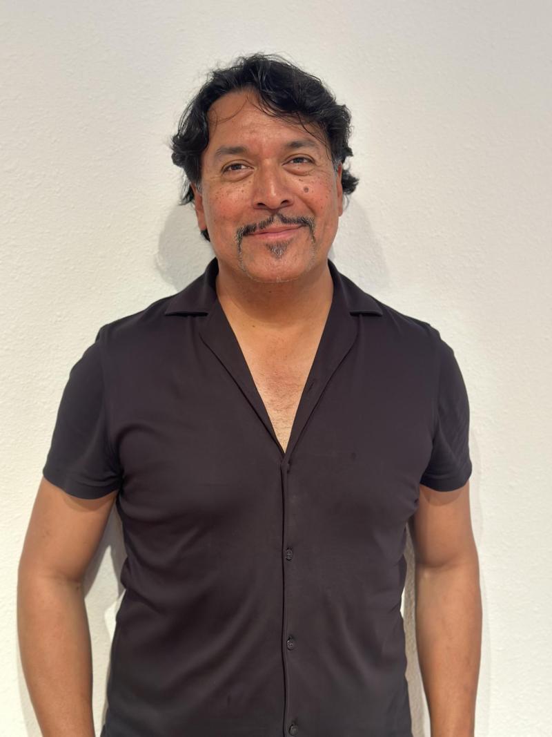 A man wearing a black shirt stands in front of a white wall.